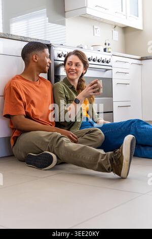Verschiedene Paare sitzen auf gefliestem Küchenboden und lehnen sich an Schränke, Frau mit Keramikbecher Stockfoto
