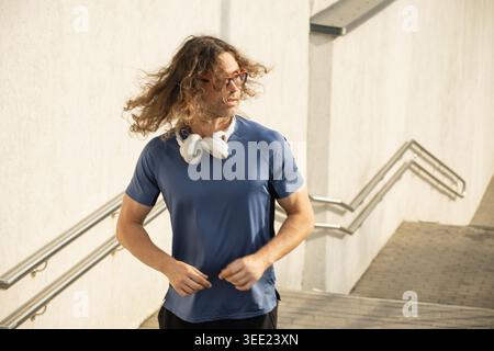 Sportlicher Läufer mit langen Haaren bei Sonnenlicht mit kabellosen Kopfhörern während des Cardio-Workouts im Freien in der Nähe von städtischen Treppen Stockfoto