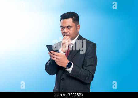 Porträt eines nachdenklichen indonesischen Führers in einem Anzug, der ein Smartphone mit der Hand am Kinn in einer denkenden Pose vor blauem Hintergrund hält. Stockfoto