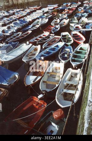 Eine bunte Auswahl an kleinen Holzfischbooten, die eng zusammen in einem ruhigen, sonnendurchfluteten Hafen in Italien der 1980er Jahre liegen Stockfoto
