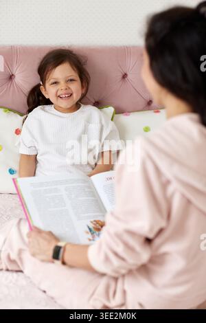 Mutter und Tochter verbinden sich, lesen zusammen im Bett zu Hause. Das Kind lächelt und genießt die Geschichtenzeit mit den Eltern Stockfoto