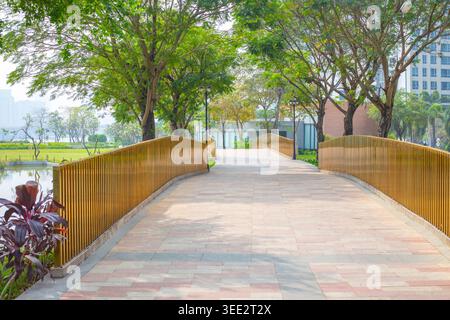 Ein sauberer Holzbrückenweg führt durch einen grünen Park mit üppigen Bäumen und Gebäuden im Hintergrund. parkweg, urbane Natur, Stadtgarten. Stockfoto