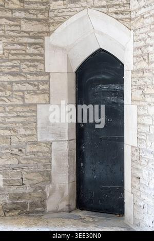 Schmaler, spitzer Bogengang mit einer verwitterten schwarzen Holztür, die in eine helle gotische Steinmauer mit strukturierten Mauerwerksblöcken eingefasst ist. Stockfoto