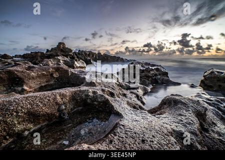 Blick auf eine zerklüftete felsige Küste mit Gezeitenbecken und Wellen, die bei Sonnenaufgang unter bewölktem Himmel gegen die Küste stürzen. Stockfoto
