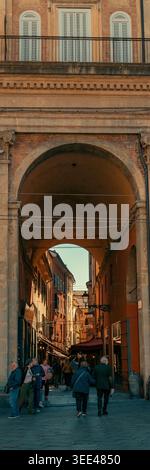 Eine vertikale Aufnahme, die einen klassischen italienischen Torbogen in Bologna, Italien, zeigt und einen Blick auf eine geschäftige, sonnendurchflutete Straße dahinter wirft. Fußgänger sind sichtbar Stockfoto