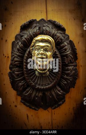 Aus nächster Nähe ein antiker goldener Türklopfer in Form eines menschlichen Kopfes, der auf einem dunklen, verzierten Holzsockel auf einem rustikalen hellbraunen hölzernen Do steht Stockfoto