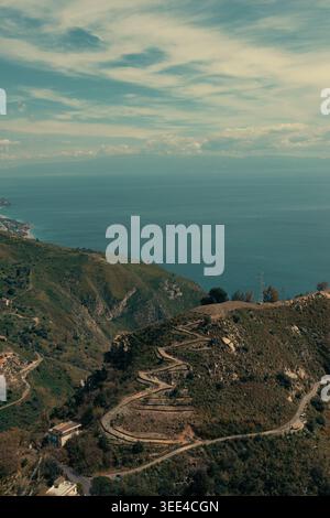 Aus der Vogelperspektive fängt die zerklüfteten, grünen Hügel von Taormina, Sizilien, ein, die hinunter zum tiefblauen Ionischen Meer führen. Kurvenreiche Straßen durchschneiden den sl Stockfoto