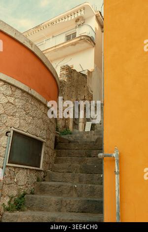 Ein nach oben gerichteter Blick durch eine enge Gasse mit alten Steintreppen, die zwischen Gebäuden hindurch führen. Ein Teil einer leuchtenden orangefarbenen Wand steht auf t Stockfoto