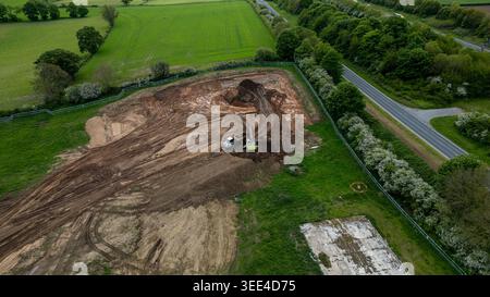Bagger und Lkw arbeiten auf einem großen, von grünen Feldern und Bäumen umgebenen, unbefestigten Gebiet in Bellway Homes in Boroughbridge, Großbritannien. Stockfoto