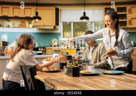 Mutter gießt Orangensaft für ihre Tochter am Frühstückstisch und serviert das Essen mit Gebäck und Waffeln oder Eiern, morgens in einer hellen Küche. Eltern und junge Mädchen essen. Stockfoto