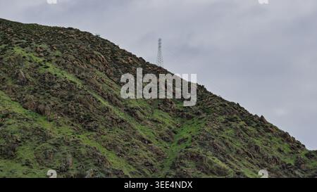 Ein hoher Stahlturm steht auf einem steilen, felsigen Kamm, der mit grünem Gras bedeckt ist Stockfoto