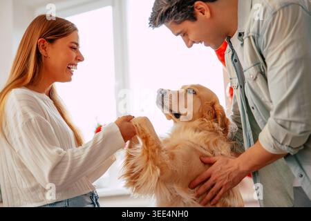 Glückliches junges Paar, das mit seinem Hund zu Hause spielt, lächelnde Tierbesitzer, die sich mit goldenem Hund in einem gemütlichen, modernen Wohnzimmer verbinden Stockfoto