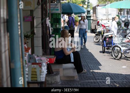 BANGKOK, THAILAND, 5. April 2026: Weibliche Touristen ruhen und Trinkwasser vor dem 24-Stunden-Geschäft, Seitenprofil, Maha Rat Road. Stockfoto