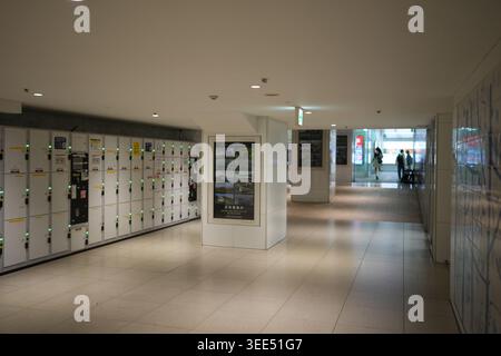 Moderner Flur mit Reihen von Schließfächern und digitalen Displays. Die Leute laufen ruhig durch einen verglasten Korridor. Helle Lichter reflektieren den polierten Boden Stockfoto