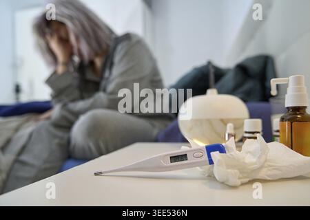 Frau im grauen Pyjama sitzt auf dem Bett mit Kopfschmerzen, Thermometer und Taschentüchern auf einem Tisch, eine Erkältung Stockfoto