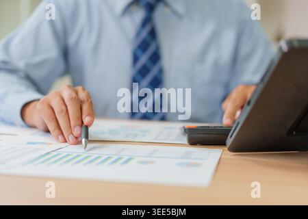 Asiatischer männlicher Profi in seinen 30ern verwendet einen Stift und Taschenrechner, um Immobilienmarktdiagramme in einem thailändischen Büro für Hypotheken und Immobilien zu analysieren Stockfoto