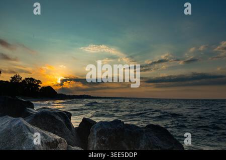 Wellen berühren sanft die Felsen entlang des Ufers, während die Sonne hinter den Wolken untergeht Bäume säumen den Strand und bilden eine natürliche Grenze, von der der Himmel Farben zeigt Stockfoto