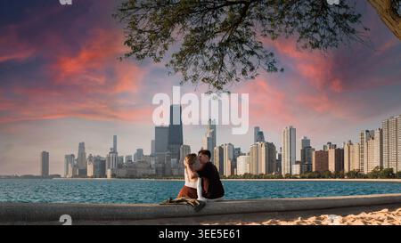 Ein Paar sitzt dicht beieinander am Seeufer in Chicago. Sie teilen sich einen Kuss, während die Sonne untergeht, und malen den Himmel mit Farben. Die Skyline von Chicag Stockfoto