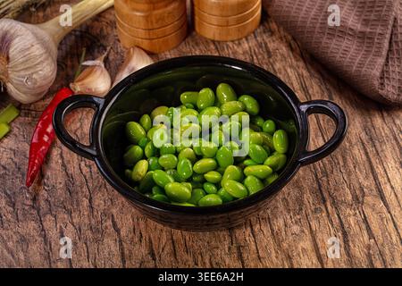 Leuchtend grüne Edamame-Bohnen in schwarzer Keramikschale, umgeben von rustikalen Kochzutaten auf strukturierter Holzoberfläche. Holzmahlmaschinen, Knoblauch, Chili Stockfoto