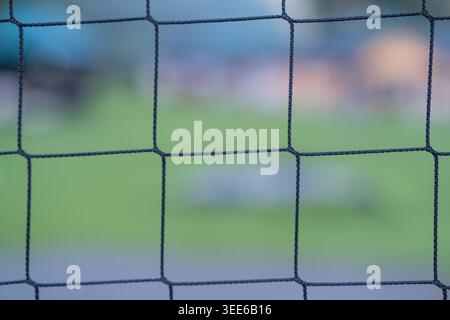 Nahaufnahme eines schwarzen Netzzaun mit verschwommenem grünem Gras und undeutlichen Objekten im Hintergrund, wodurch ein weicher Fokus entsteht Stockfoto
