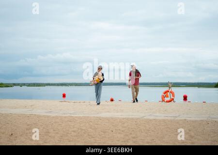 Ein Mann und eine Frau schlendern friedlich an einem ruhigen Strandufer am See entlang. Der Mann und seine Frau genießen einen ruhigen, romantischen Spaziergang im Freien. Partner teilen einen ruhigen Moment am Sandstrand Stockfoto