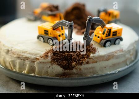 Hausgemachte Baugeburtstorte mit Spielzeugbagger und Müllwagen auf weißem Frosting. Schokoladenbrösel als Schmutz und Sand auf der Baustelle. Kreatives Dessert für Kinder, zubereitet von Mutter Stockfoto