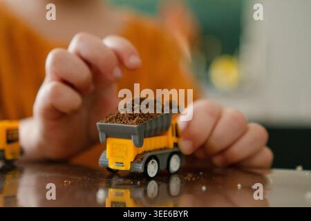 Kinderhände tragen einen gelben Kipper mit Erde über eine reflektierende Oberfläche. Fantasievolle Kinder bauen kreative Spielwelten mit Miniatur-Fahrzeug auf. Ein fokussierter Youngster baut drinnen eine Fantasieszene auf Stockfoto