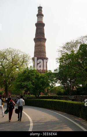 Blick auf das historische Minarett Qutub Minar in Delhi, Indien, mit Besuchern, die auf einem gepflasterten Pfad zwischen grünen Bäumen spazieren Stockfoto