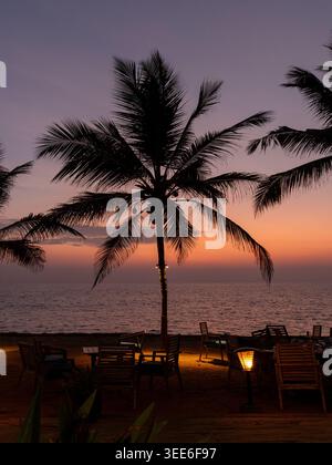 Silhouette von Kokospalmen und Restauranttischen am Strand mit Laterne in der Dämmerung über dem Arabischen Meer, Kerala Küste, Indien Stockfoto