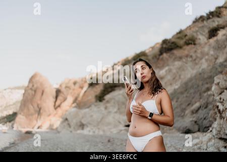 Frau, Strand, Telefon. Junge Frau in weißem Bikini mit Smartphone am felsigen Sommerstrand mit Kopierraum. Stockfoto