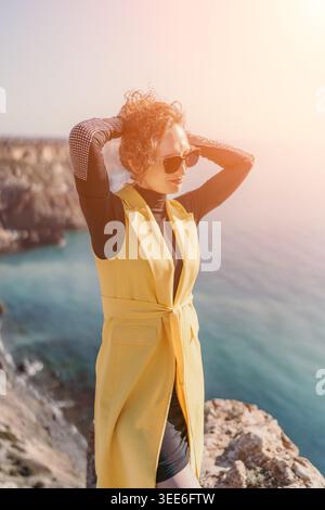 Frau Klippe Ozean Frau in gelber Weste steht an der felsigen Küste mit Blick auf das Meer während Sonnenuntergang goldene Stunde Stockfoto