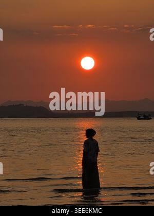 Silhouette einer Person, die im Wasser steht und den hellen orangefarbenen Sonnenuntergang über dem Ozean beobachtet und sich friedlich und friedlich anfühlt Natur Schönheit ruhige Meer dista zu betrachten Stockfoto
