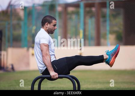 Sportlicher Mann beim L-Sit auf parallelen Bars in einem Fitnessstudio im Freien, konzentriert sich auf Kraft, Calisthenics und Crossfit-Training mit einem Freund, zeigt Balance, Core Control und disziplinierte Workout-Form. Stockfoto