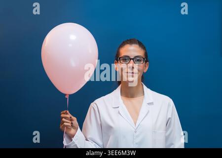 Weibliche Ärztin und Labortechnikerin in einem weißen Mantel hält einen rosa Ballon vor einem blauen Hintergrund und vermittelt Leichtigkeit, Selbstvertrauen und einen Moment der Feier in einem klinischen Labor. Stockfoto