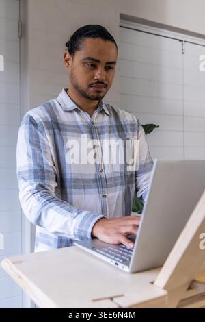 Asiatischer Mann tippt auf silbernem Laptop, steht am Schreibtisch und trägt kariertes Hemd im Büro mit Pflanze. Einstellbar, Staffelei, Workstation, minimalistisch, neutral, Grün, Tageslicht Stockfoto