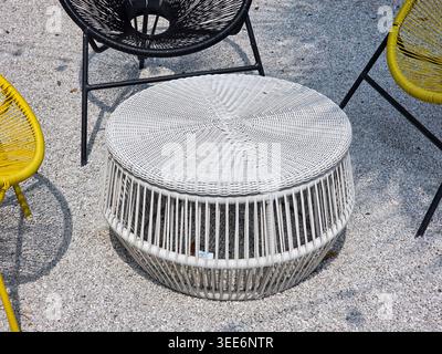 Der weiße, gewebte Korb-Couchtisch im Freien befindet sich auf einer Betonterrasse, umgeben von farbenfrohen Lounge-Stühlen im Retro-acapulco-Stil, die entspannend einladend wirken Stockfoto