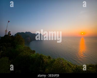 Friedliche Person sitzt auf einer hohen grünen Klippe und beobachtet den wunderschönen orangefarbenen Sonnenuntergang über ruhigem Ozeanwasser. Es fühlt sich ruhig und ruhig an und beobachtet den fernen Horizont Stockfoto