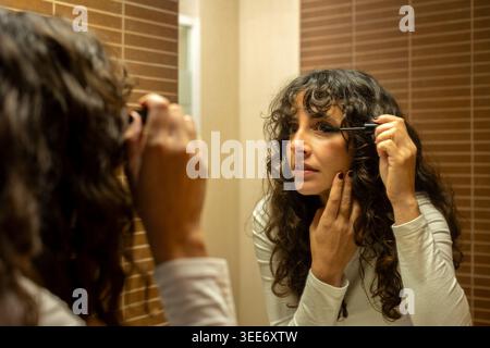 Eine junge Frau, die Mascara aufträgt, während sie sich im Badezimmerspiegel ansieht. Authentisches, benutzergeneriertes Inhaltsbild mit einem echten täglichen Make-up Stockfoto