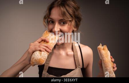 Junge Frau genießt es, in frisches knuspriges Brotbaguette zu beißen, mit Schürze und lächelndem Betrachter Stockfoto