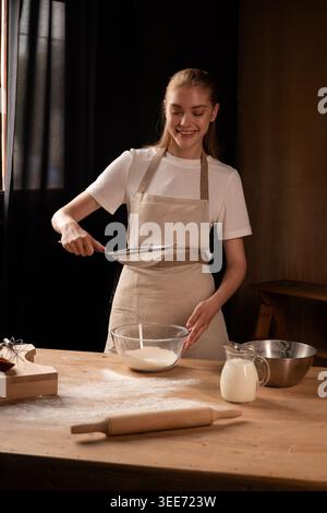 Lächelnde Frau siebt Mehl in eine Schüssel auf einem Holztisch und bereitet Zutaten zum Backen vor Stockfoto