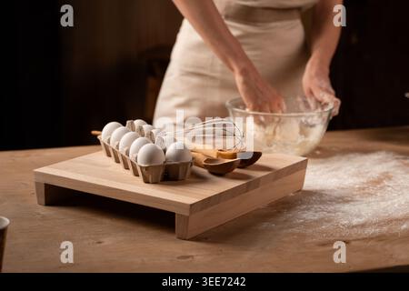Baker kneten Teig in einer Glasschale mit Eiern, Schneebesen und Holzutensilien auf einem rustikalen Küchentisch Stockfoto