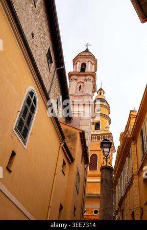 Niedriger Blick auf die gelben Glockentürme der Basilika von Menton, die sich über den engen Altstadtstraßen an der französischen Riviera erheben Stockfoto