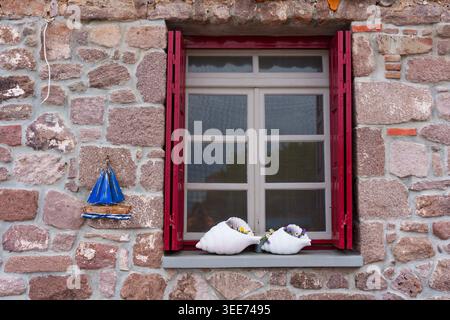Rustikales Steinhausfenster in Skala Sikamineas, Griechenland, mit roten Rollläden, einem dekorativen blauen Segelboot und großen weißen Muschelpflanzen auf der si Stockfoto