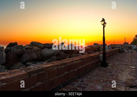 Blick auf den Sonnenuntergang über die Steinmauer des Hafens und die alte Straßenlaterne in Molyvos, Lesbos, Griechenland. Leuchtender orangefarbener Himmel hebt die Küstenlandschaft in der Abenddämmerung hervor Stockfoto