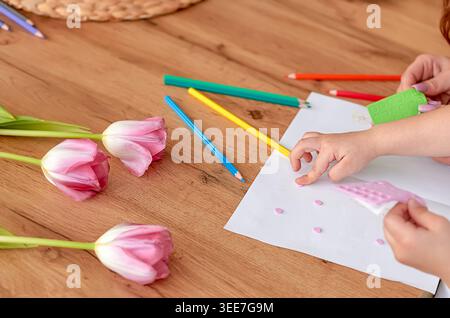 Kinderhände verwenden farbige Bleistifte und Bastelmaterialien auf einem Holztisch, umgeben von rosa Tulpen und einem leeren Blatt Papier Stockfoto