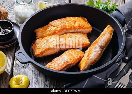 Gebratene Lachsfischfilets in einer gusseisernen Pfanne mit frischen Zitronenscheiben, Meersalz, schwarzem Pfeffer und Koriander auf einem rustikalen Holztisch, horizontale Ansicht, Stockfoto