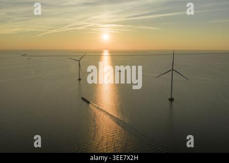 Luftaufnahme von Offshore-Windturbinen und einem Frachtschiff, das während eines goldenen Sonnenuntergangs in den Niederlanden auf dem ruhigen Meer segelt. Stockfoto