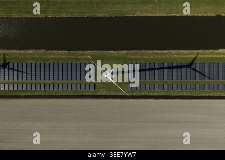Luftaufnahme einer Windkraftanlage, die einen langen Schatten über Reihen von Solarpaneelen neben einem Kanal und landwirtschaftlichen Feldern in den Niederlanden wirft. Stockfoto