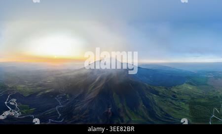 Aus der Vogelperspektive auf den Ätna mit Dampf, der aus dem Krater steigt, und gewundenen Straßen an den Vulkanhängen bei Sonnenuntergang Castiglione di Sicilia, Sizilien, Italien. Stockfoto