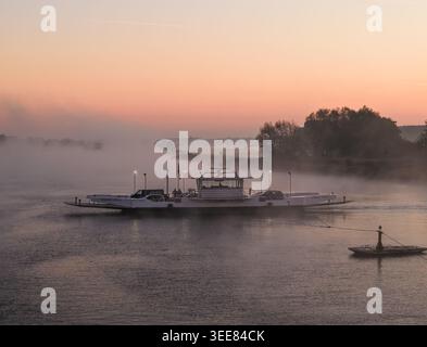 Aus der Vogelperspektive einer Autofähre, die einen nebeligen Fluss überquert, während eines sanften orangen Sonnenaufgangs mit dickem Nebel über dem Wasser und weit entfernten Bäumen Kessel, Limburg Stockfoto